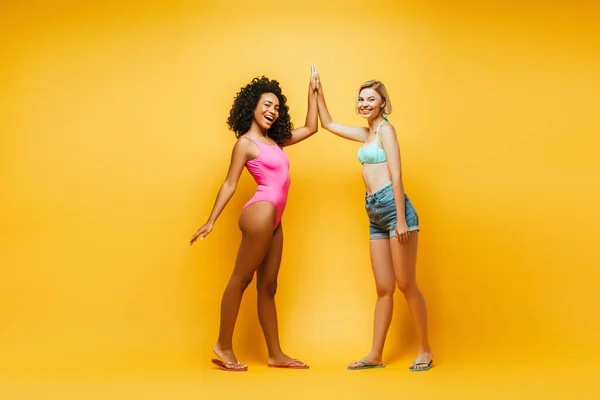 Vue pleine longueur de la jeune femme afro-américaine en maillot de bain donnant haute cinq à un ami blonde en short sur jaune — Photo de stock