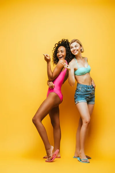 Visão de comprimento completo de rir mulheres interracial na roupa de verão apontando com os dedos e olhando para longe em amarelo — Fotografia de Stock