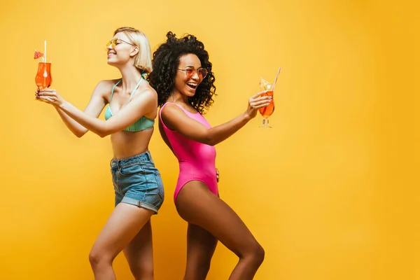 Jóvenes mujeres multiculturales en traje de verano de pie espalda con espalda con copas de cóctel en amarillo - foto de stock