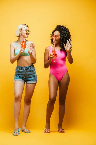 Young african american woman showing hello gesture near blonde friend while holding cocktail glasses  on yellow — Stock Photo