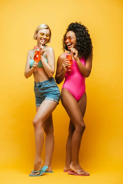 Mujer rubia en pantalones cortos y amigo afroamericano en traje de baño con copas de cóctel en amarillo - foto de stock