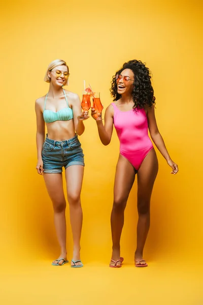 Vista completa de la mujer afroamericana en traje de baño tintineo copas de cóctel con amigo rubio en amarillo - foto de stock