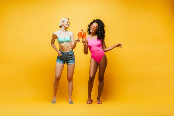 Полноразмерный вид блондинки в шортах, звенящей в коктейльных очках с африканским американским другом в купальнике на желтом — стоковое фото