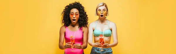 Horizontal image of shocked interracial women in summer outfit holding cocktails and looking at camera isolated on yellow — Stock Photo