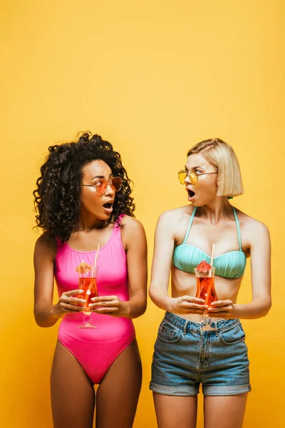 Шоковані міжрасові друзі в літньому вбранні тримають коктейлі і дивляться один на одного на жовтому — стокове фото