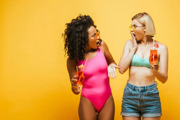 Shocked interracial women in summer outfit with cocktail glasses looking at each other on yellow — Stock Photo