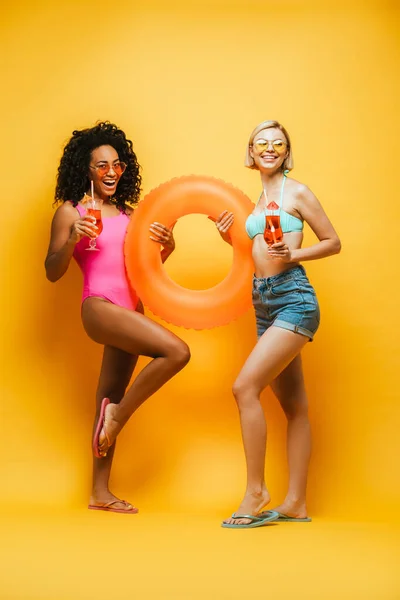 Visão completa do comprimento de mulheres multiculturais excitadas na roupa do verão que prende o anel inflável e os copos de coquetel no amarelo — Fotografia de Stock