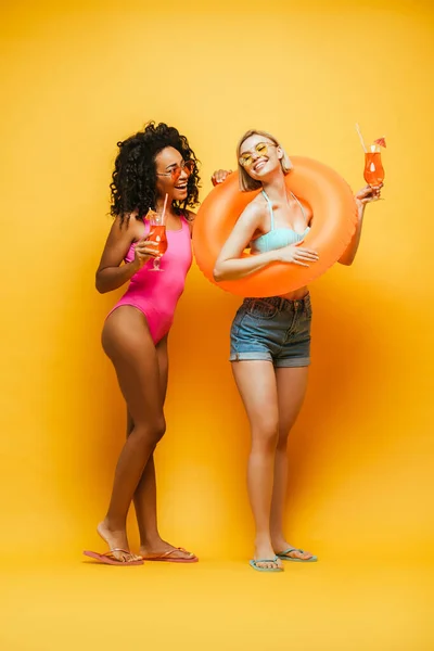 Joven afroamericana mujer en traje de baño mirando rubia amiga con anillo de baño en amarillo — Stock Photo