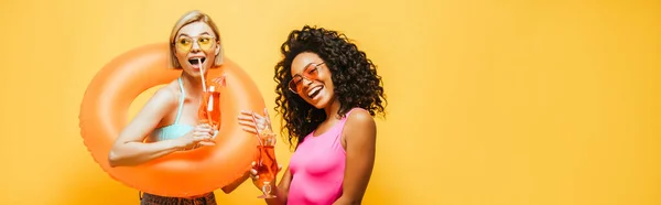 Imagen horizontal de mujeres jóvenes interracial con anillo de baño y vasos de cóctel posando aislados en amarillo - foto de stock