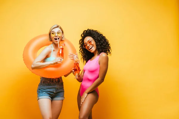 Emocionadas mujeres interracial con anillo de natación y copas de cóctel posando en amarillo — Stock Photo