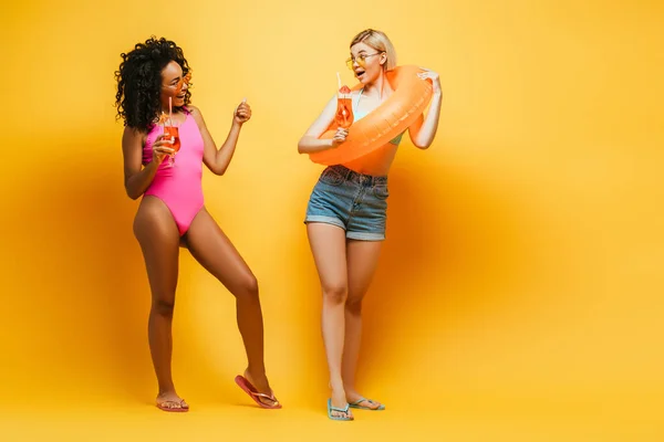 Young african american woman in swimsuit, and blonde friend with swim ring holding cocktail glasses on yellow — Stock Photo