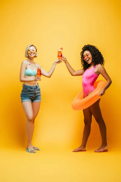 Excited interracial women in summer outfit clinking cocktail glasses and looking at camera on yellow — Stock Photo