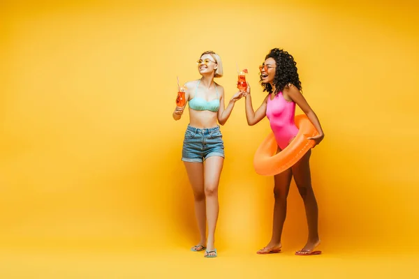 Mulher loira com amigo americano africano segurando copos de coquetel e anel de natação rindo enquanto olha para o amarelo — Fotografia de Stock