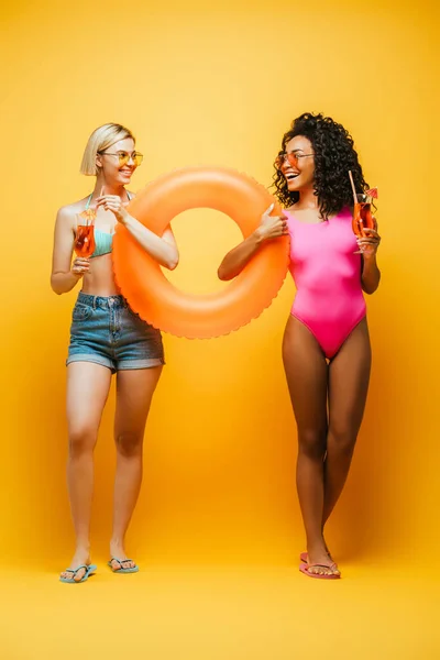 Junge interrassische Frauen im Sommer-Outfit mit Schwimmring und Cocktailgläsern, während sie sich auf gelb anschauen — Stockfoto