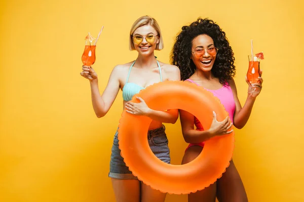 Jóvenes mujeres interracial en traje de verano celebración de anillo de natación y vasos de cóctel en amarillo - foto de stock