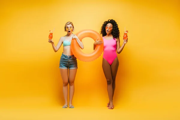 Surprised blonde woman in shorts with african american friend in swimsuit holding swim ring and cocktail glasses on yellow — Stock Photo