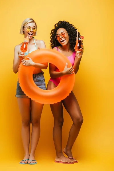 Jóvenes mujeres multiculturales en traje de verano posando con anillo inflable y cócteles en amarillo — Stock Photo