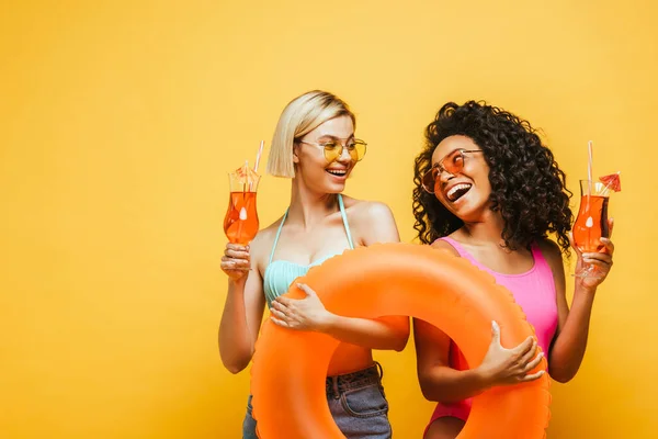 Excitadas mulheres inter-raciais com anel de natação e coquetel olhando um para o outro em amarelo — Fotografia de Stock