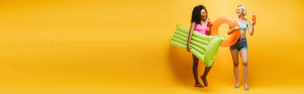 Imagem horizontal de mulheres jovens multiétnicas em roupa de verão posando com colchão inflável, anel de natação e coquetéis em amarelo — Fotografia de Stock