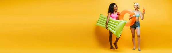 Horizontal image of multiethnic young women in summer outfit posing with inflatable mattress, swim ring and cocktails on yellow — Stock Photo