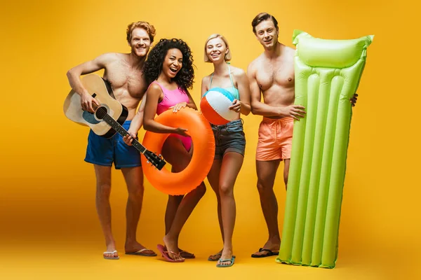 Full length view of excited multicultural friends with guitar, inflatable mattress, ball and swim ring looking at camera on yellow — Stock Photo