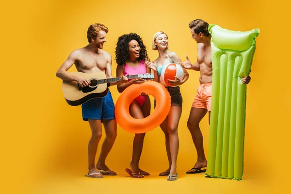Visão completa do comprimento de amigos multiculturais excitados com guitarra, colchão inflável, bola e anel de natação falando no amarelo — Fotografia de Stock