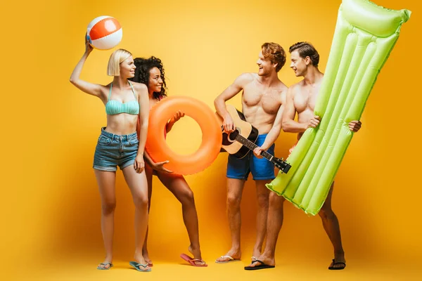 Visão de comprimento total de jovens amigos multiculturais em roupa de verão com guitarra, colchão inflável, bola e anel de natação olhando um para o outro em amarelo — Fotografia de Stock