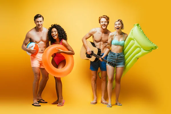 Vista completa de amigos jóvenes multiculturales con guitarra, bola inflable, colchón y anillo de natación mirando a la cámara en amarillo - foto de stock