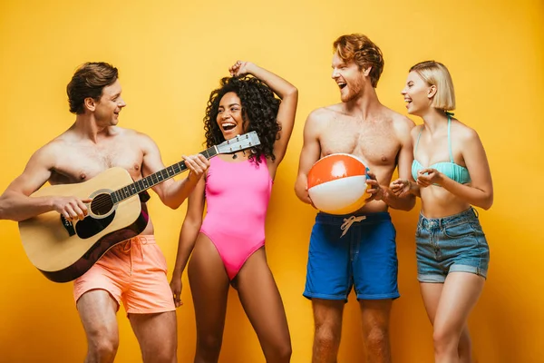 Jovem tocando guitarra acústica perto de amigos multiétnicos animados em roupa de verão em amarelo — Fotografia de Stock