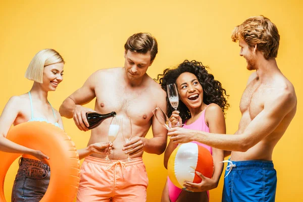 Young man pouring champagne in glasses of multicultural friends holding inflatable ball and ring isolated on yellow — Stock Photo