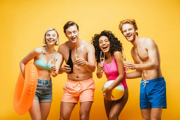 Amici multiculturali eccitati con bicchieri di champagne, anello gonfiabile e palla guardando la fotocamera isolata sul giallo — Foto stock
