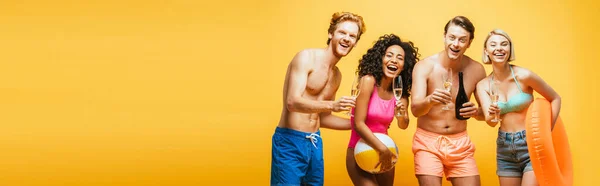 Imagen horizontal de amigos multiculturales excitados sosteniendo la bola inflable, anillo de natación y vasos de cóctel aislados en amarillo - foto de stock