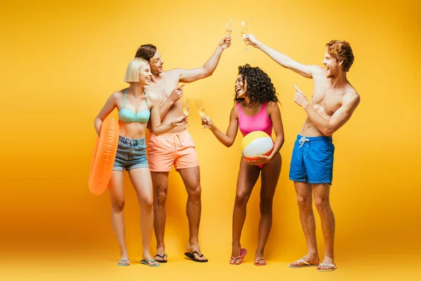Full length view of shirtless man clinking champagne glasses near multicultural women with inflatable ball and ring on yellow — Stock Photo