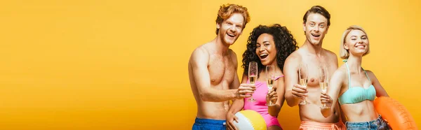 Imagem horizontal de jovens amigos multiculturais segurando copos de champanhe, anel inflável e bola enquanto olha para a câmera isolada no amarelo — Fotografia de Stock