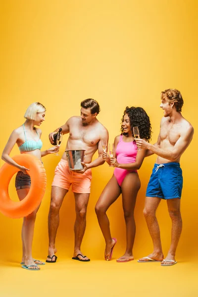 Full length view of man pouring champagne in glasses of multicultural friends wearing summer outfit on yellow — Stock Photo