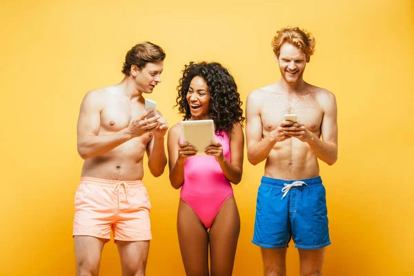 Excited multicultural friends in summer outfit using digital devices isolated on yellow — Stock Photo