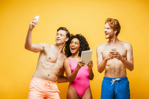 Joven tomando selfie en smartphone con amigos multiétnicos sosteniendo dispositivos digitales aislados en amarillo - foto de stock