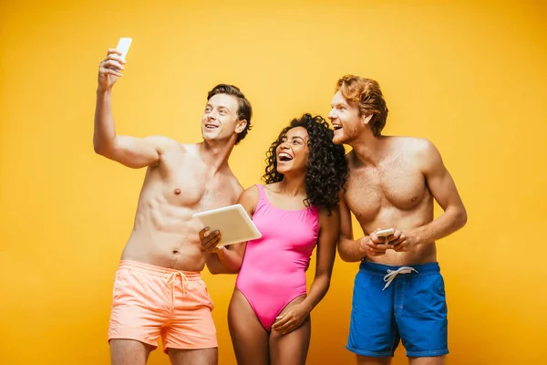 Uomo senza maglietta prendendo selfie su smartphone con eccitati amici multiculturali in possesso di gadget isolati su giallo — Foto stock