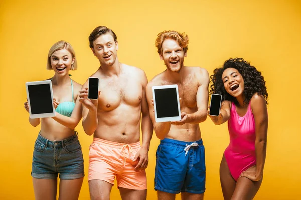 Jovens amigos multiétnicos em roupa de verão mostrando dispositivos digitais com tela em branco enquanto olha para a câmera isolada no amarelo — Fotografia de Stock