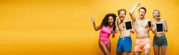 Horizontal image of excited multicultural friends in summer outfit showing gadgets with blank screen on yellow — Stock Photo