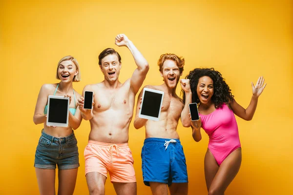 Amigos multiétnicos animado em roupa de verão mostrando dispositivos digitais com tela em branco no amarelo — Fotografia de Stock