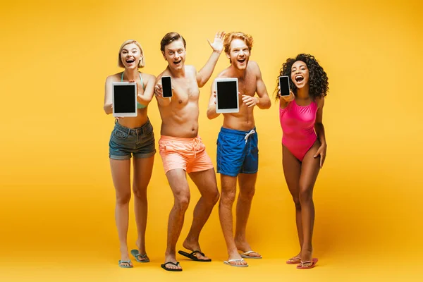Visão de comprimento total de jovens amigos multiétnicos na roupa de verão mostrando gadgets com tela em branco no amarelo — Fotografia de Stock