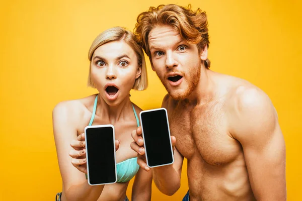 Sorprendió a la joven pareja mostrando teléfonos inteligentes con pantalla en blanco mientras miraba a la cámara aislada en amarillo - foto de stock