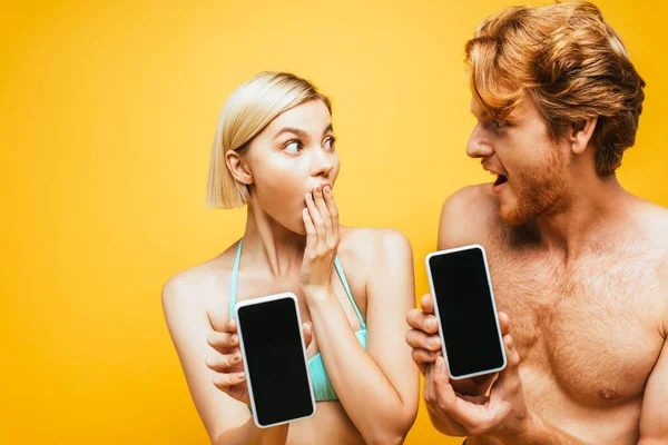 Impactó a la mujer rubia y al hombre sin camisa mostrando teléfonos inteligentes con pantalla en blanco mientras se miraban aislados en amarillo - foto de stock