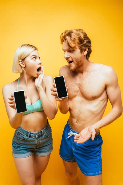 Шокована молода пара в літньому вбранні, що показує смартфони з порожнім екраном і дивиться один на одного на жовтий — стокове фото