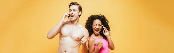 Imagen horizontal de pareja interracial excitada mostrando el gesto ganador mientras habla en teléfonos inteligentes aislados en amarillo - foto de stock