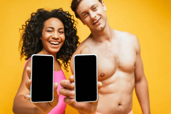 Enfoque selectivo de hombre sin camisa y mujer afroamericana mostrando teléfonos inteligentes con pantalla en blanco aislado en amarillo - foto de stock