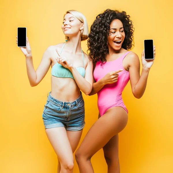 Mujer rubia con amigo afroamericano en traje de verano apuntando a teléfonos inteligentes con pantalla en blanco en amarillo - foto de stock