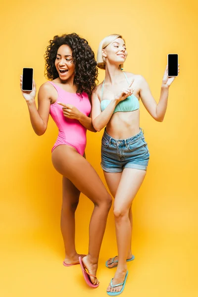 Full length view of young multicultural women in summer outfit pointing at smartphones with blank screen on yellow — Stock Photo