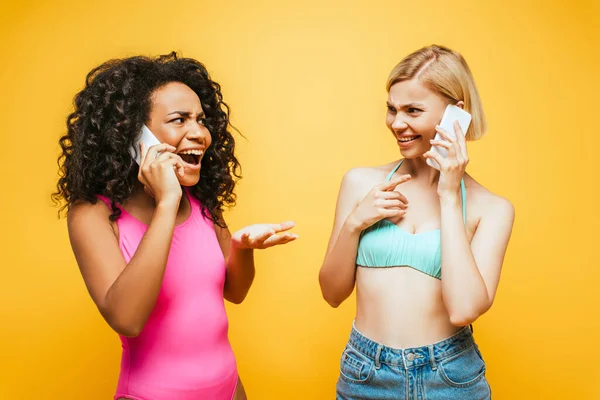 Збуджена афро-американка з блондинкою подруга говорить на смартфонах ізольовано на жовтому — стокове фото